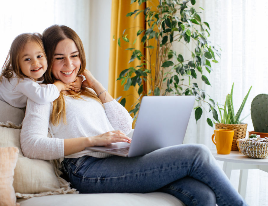 Young, happy mother laughing with daughter while sitting on the sofa cuddling and using laptop to search for dating tips for single moms.