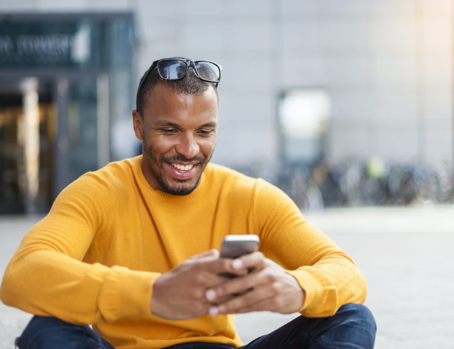 Smiling man wearing yellow pullover looking at cell phone while texting a woman that he likes.