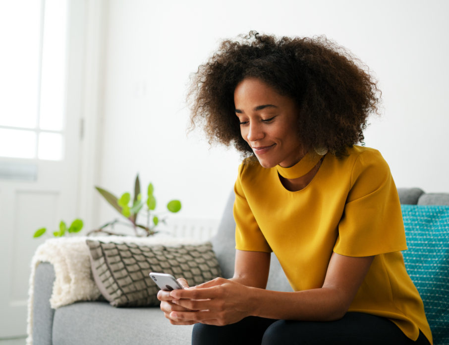 Young, smiling woman sitting on the couch using smart phone while choosing between some of the best dating apps in 2022.