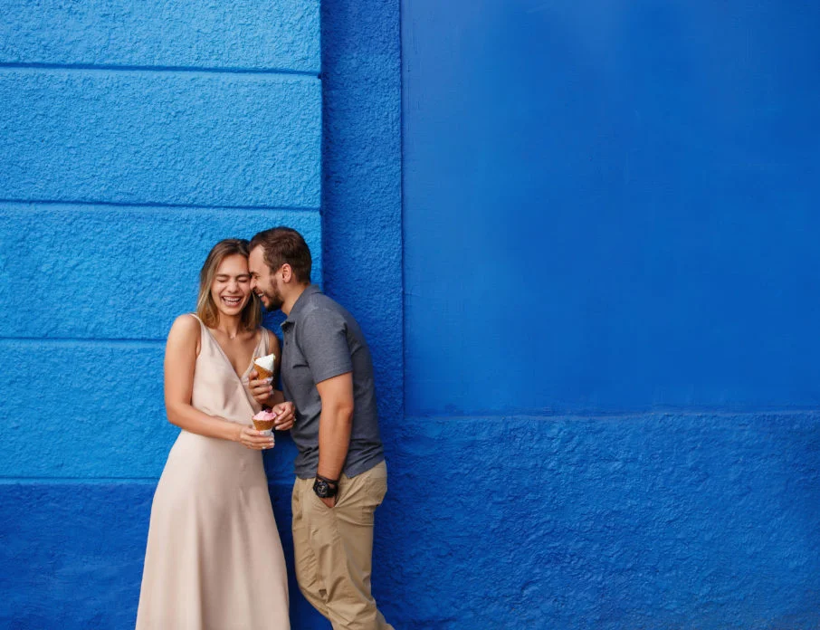 Happy, laughing man and woman standing together in front of a blue-painted wall while the woman tries out some flirty questions to ask a guy.