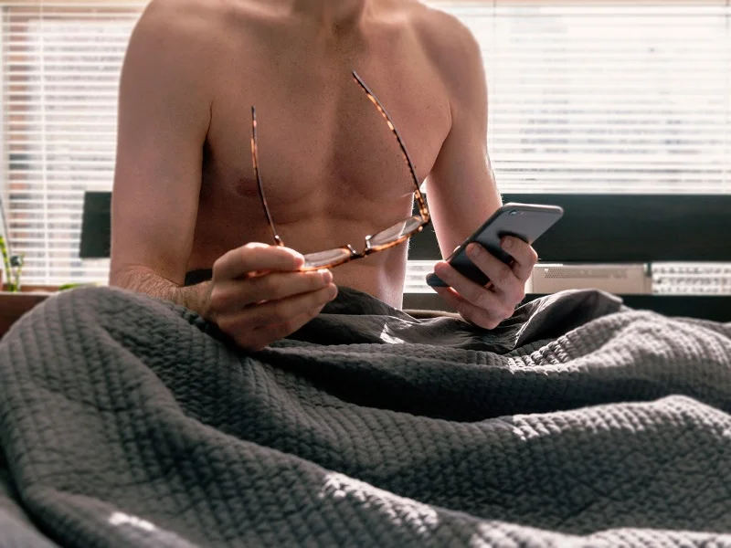 man sitting topless in bed holding a phone having a sexting chat