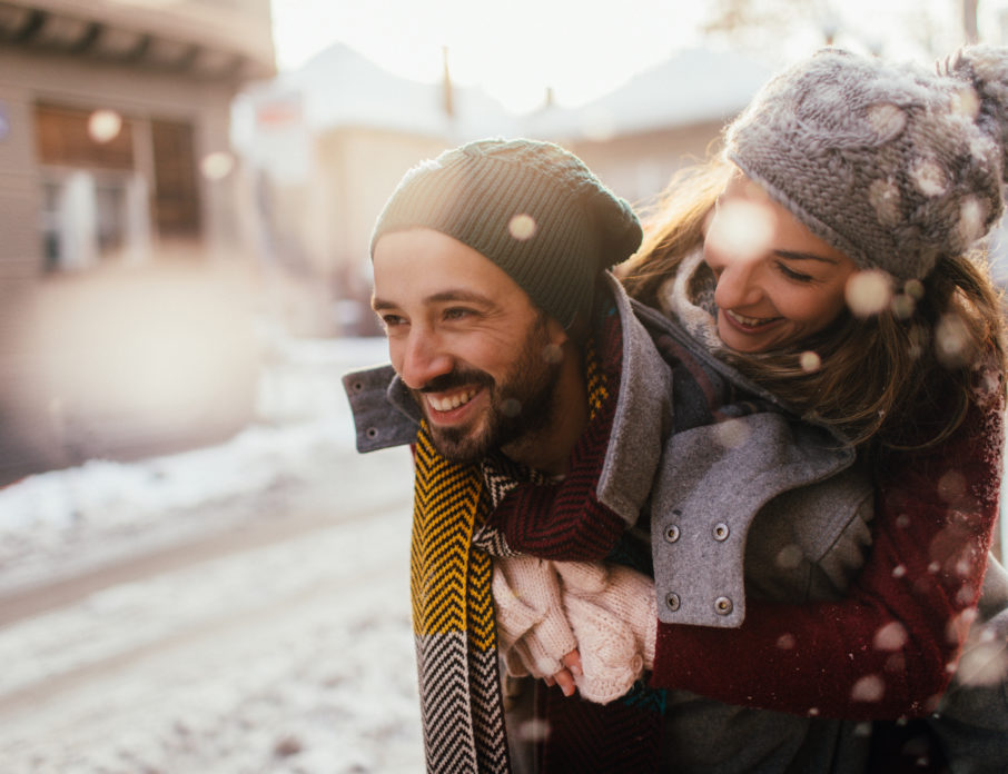 Romantic couple smiling and laughing together on a snowy day while trying out some cute winter date ideas.