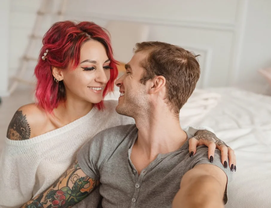Man and woman sitting on a bed in a bright room smiling at each other.