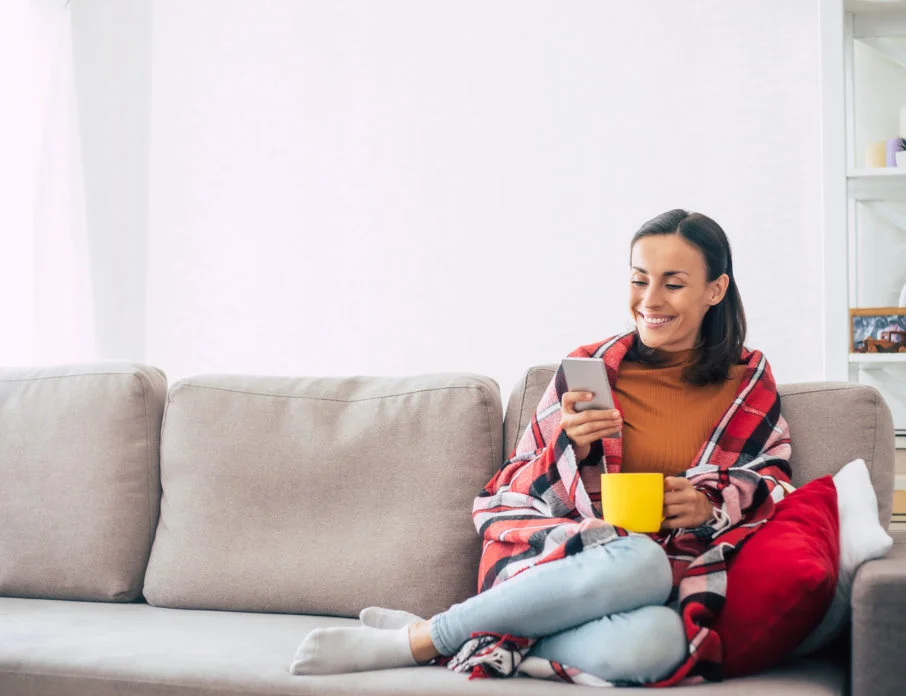 Happy woman sitting on the sofa and smiling while using her smartphone.