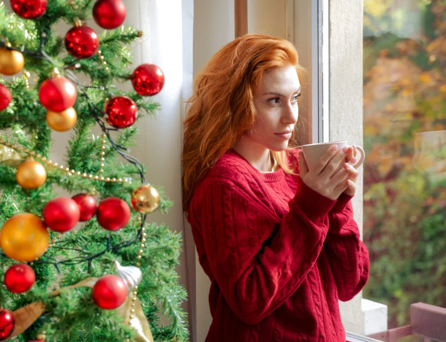 Pensive young woman standing by Christmas tree looking out of the window and thinking about how to survive the holidays after a breakup.