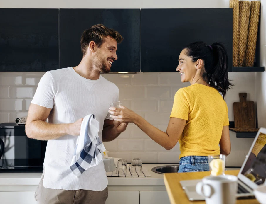 Happy young couple smiling at each while washing dishes and navigating a love-hate relationship.