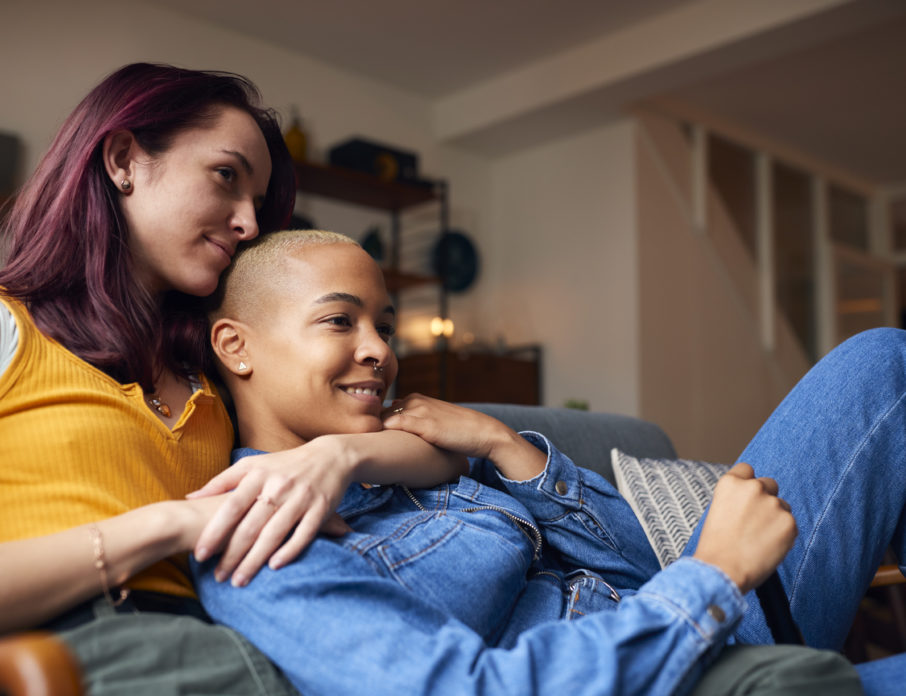 Happy, smiling lesbian couple sitting on sofa and watching some of the best LGBTQ movies on Netflix.
