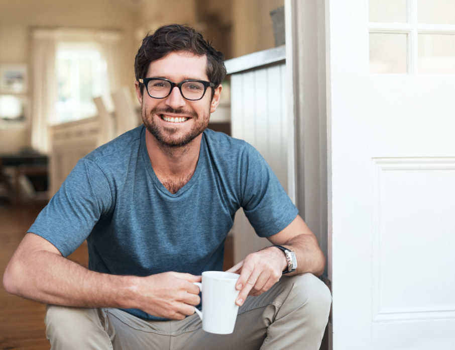 Happy, young man sitting down, holding a cup of coffee and smiling after practicing self-love for men.