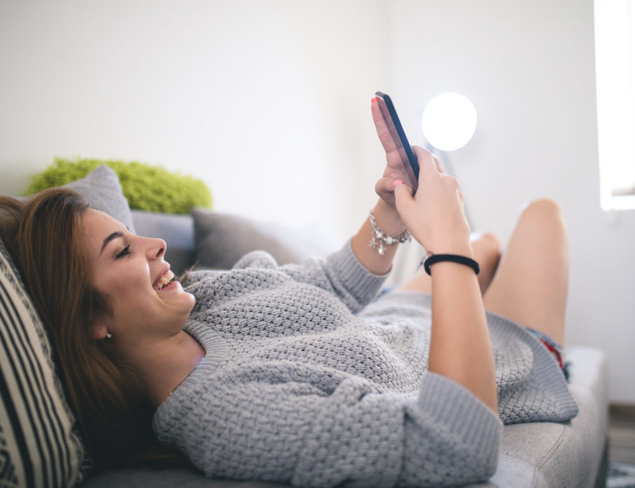 Young, smiling woman lying on sofa and using cellphone for online dating in 2022.