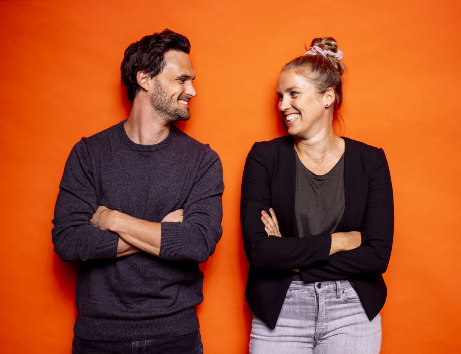 Happy, millennial dating couple smiling and looking into each other's eyes while standing in front of an orange background.
