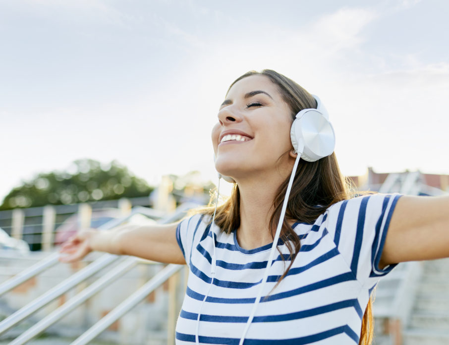 Happy, smiling woman wearing headphones and standing outside with her arms outstretched while listening to some of the best self-love songs.