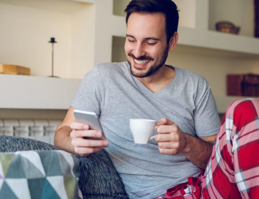 Smiling man sitting on the sofa and ready a good morning text for him while holding a cup of coffee.