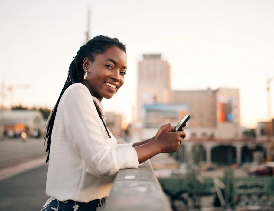 Young, smiling woman standing outdoors on a bridge and using mobile phone for Instagram dating.