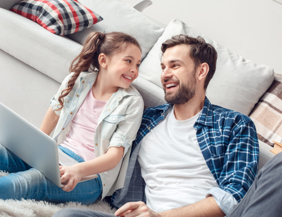 Father sitting in front of the sofa laughing with his daughter and using a laptop while dating as a single dad.