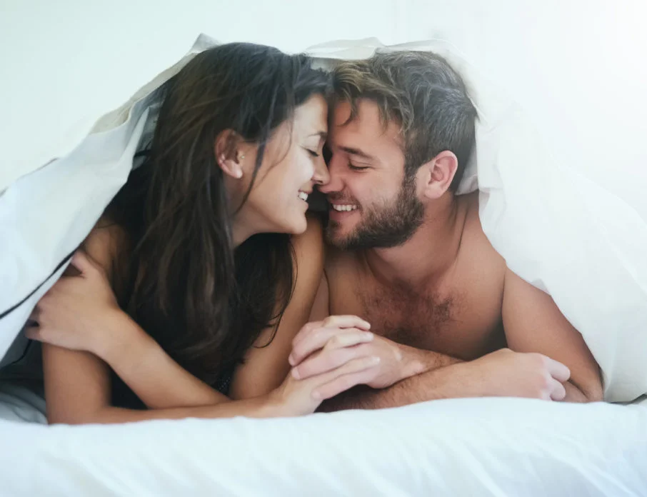 Happy man and woman lying in bed together underneath the duvet and holding hands after enjoying the health benefits of sex.
