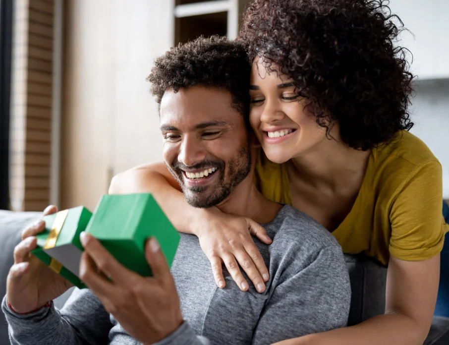 Smiling man with the gifts love language, sitting on the sofa opening a gift while girlfriend hugs him.