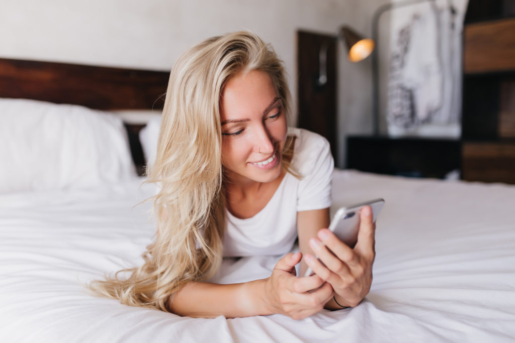 Happy woman smiling, lying on bed on her phone while online dating in Jacksonville FL.