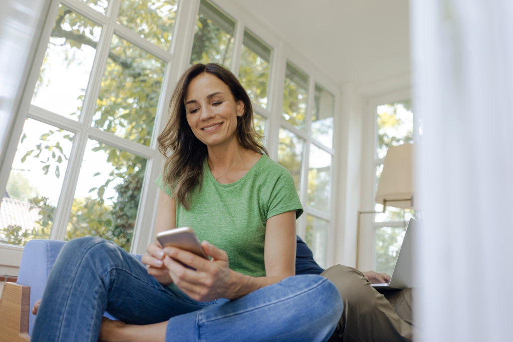 Smiling mature woman sitting on couch at home using cell phone while online dating.