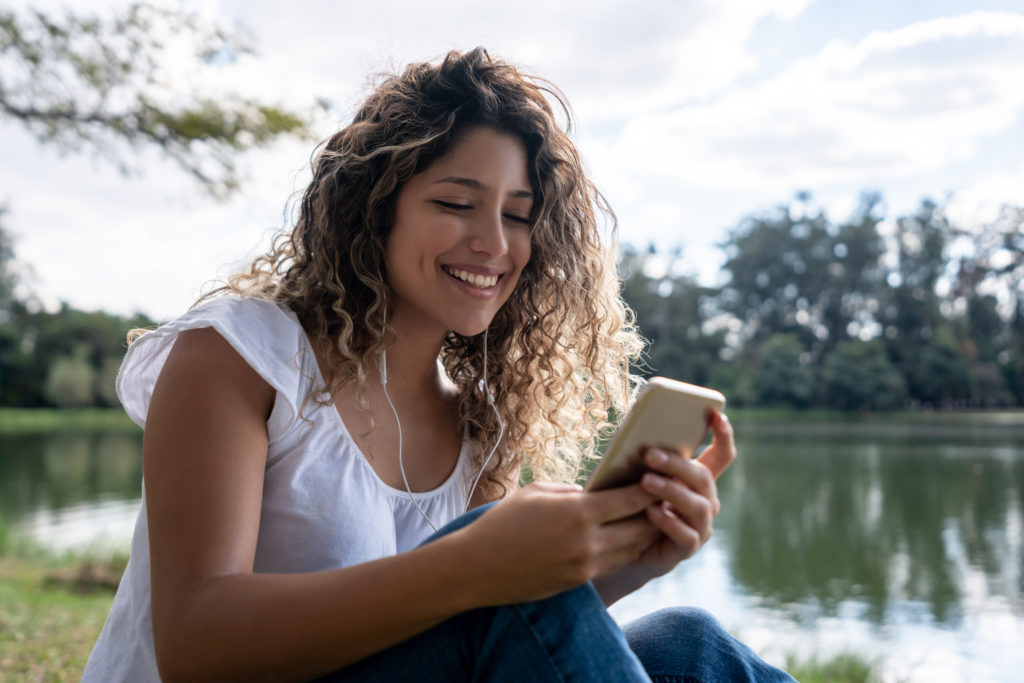 Young, smiling woman sitting outdoors with wearing earphones and using her mobile phone while dating in Tucson.