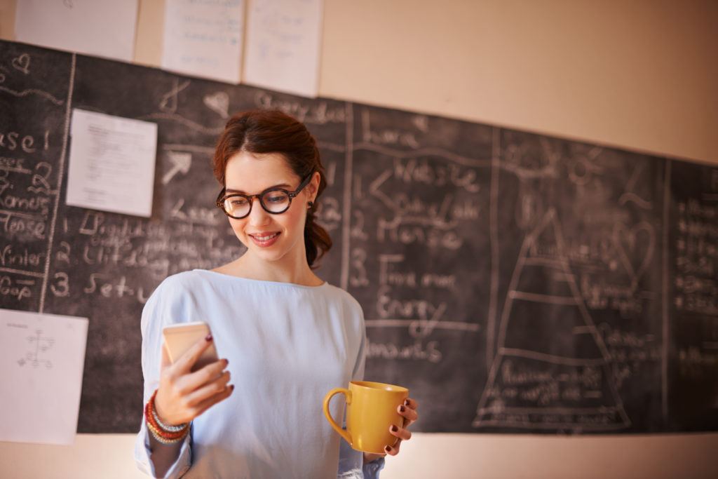 Young, smiling woman standing in her classroom and using mobile phone for teacher dating.