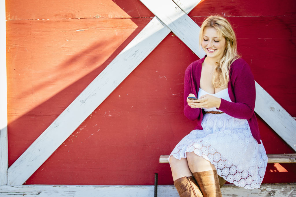 Smiling, attractive single woman sitting down on bench in front of a barn and using mobile phone to meet other Omaha singles online.