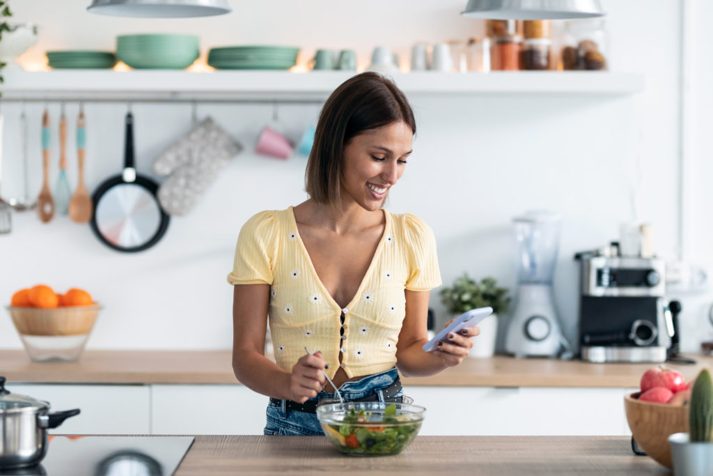Young, smiling woman standing in a kitchen preparing a salad and using her smartphone to discover the best vegan dating apps.