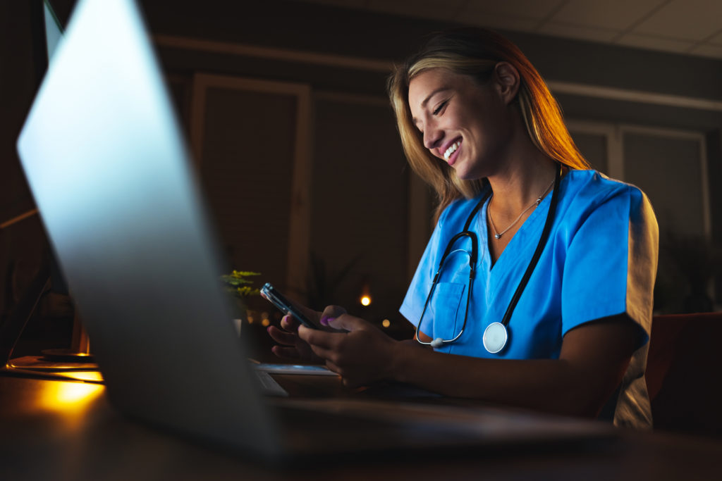 Young female nurse sitting and desk and smiling while using mobile phone to find other single nurses.