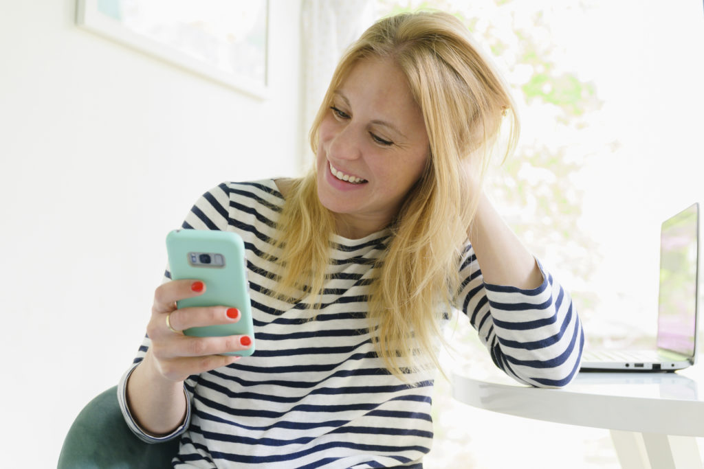 Middle-aged woman sitting at a table and smiling while looking at spiritual dating app on her mobile phone.