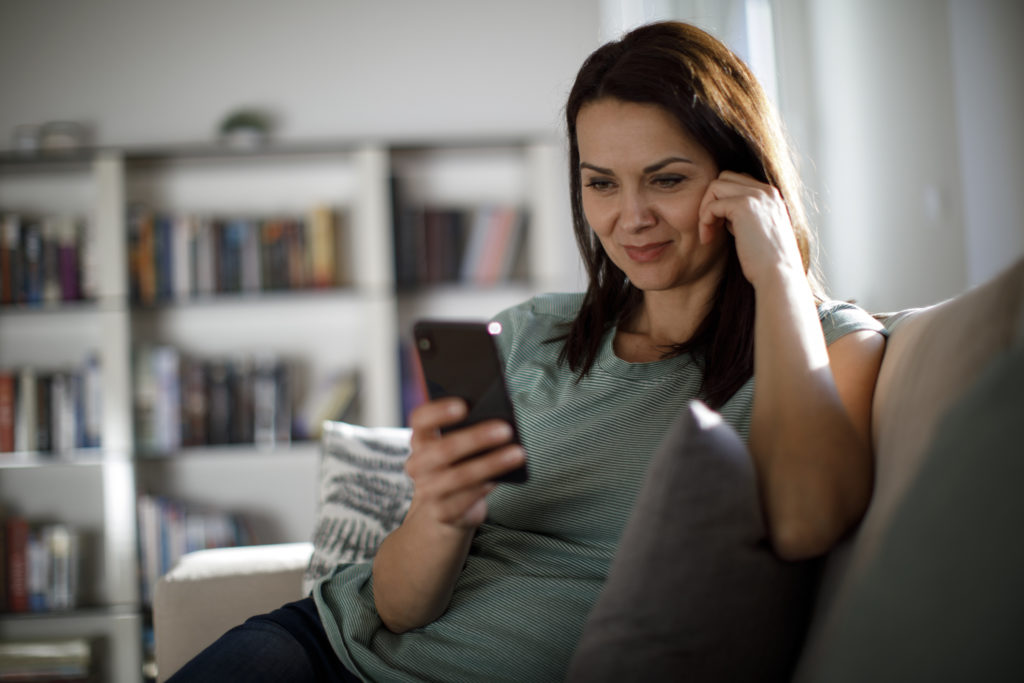 Smiling woman sitting on the the sofa and using mobile phone while online dating in Pittsburgh.