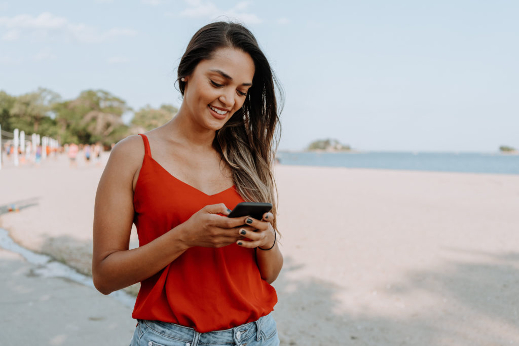 Smiling Latin single woman standing on the beach and using mobile phone for Latin dating.