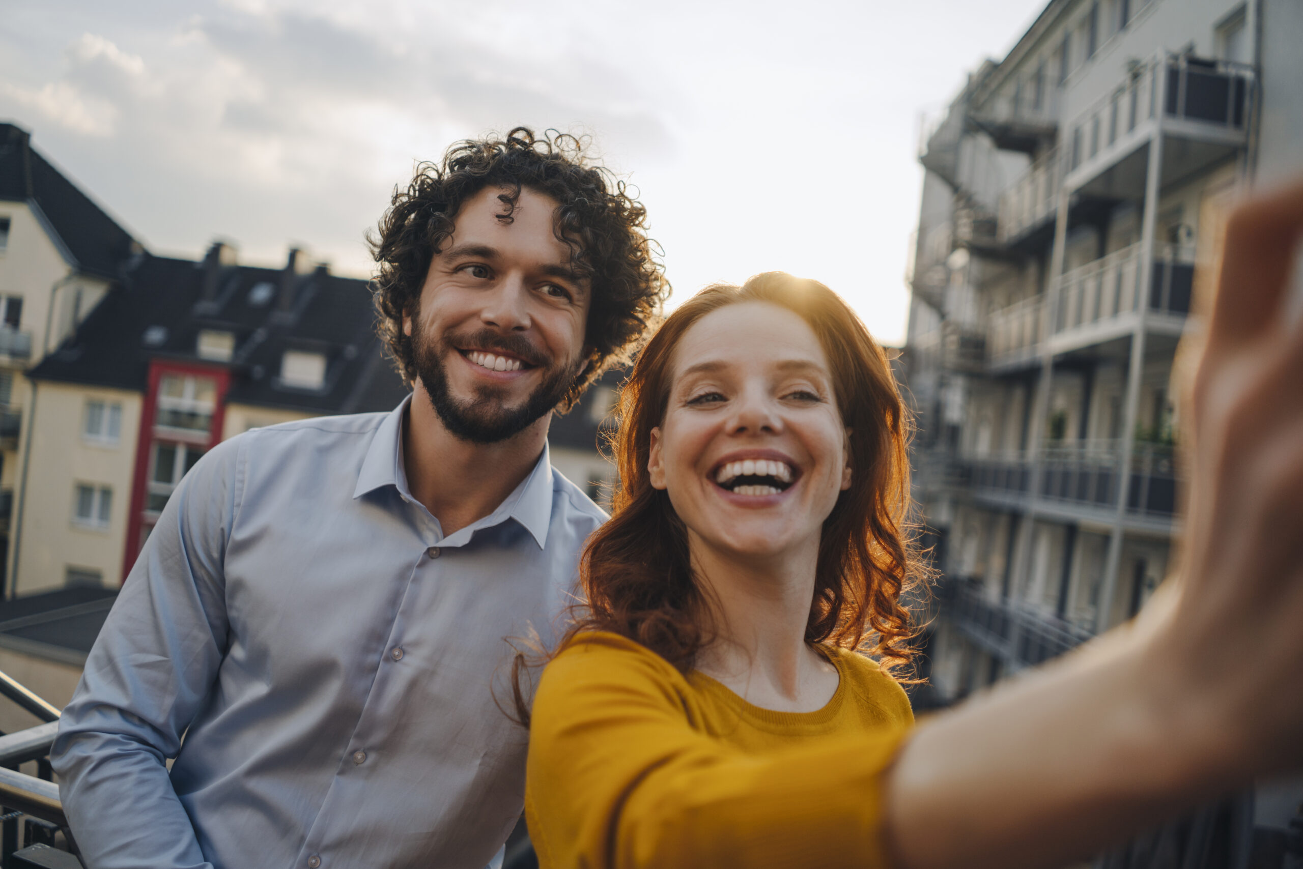 Happy couple standing outdoors taking a selfie and laughing after trying out some boyfriend nicknames.