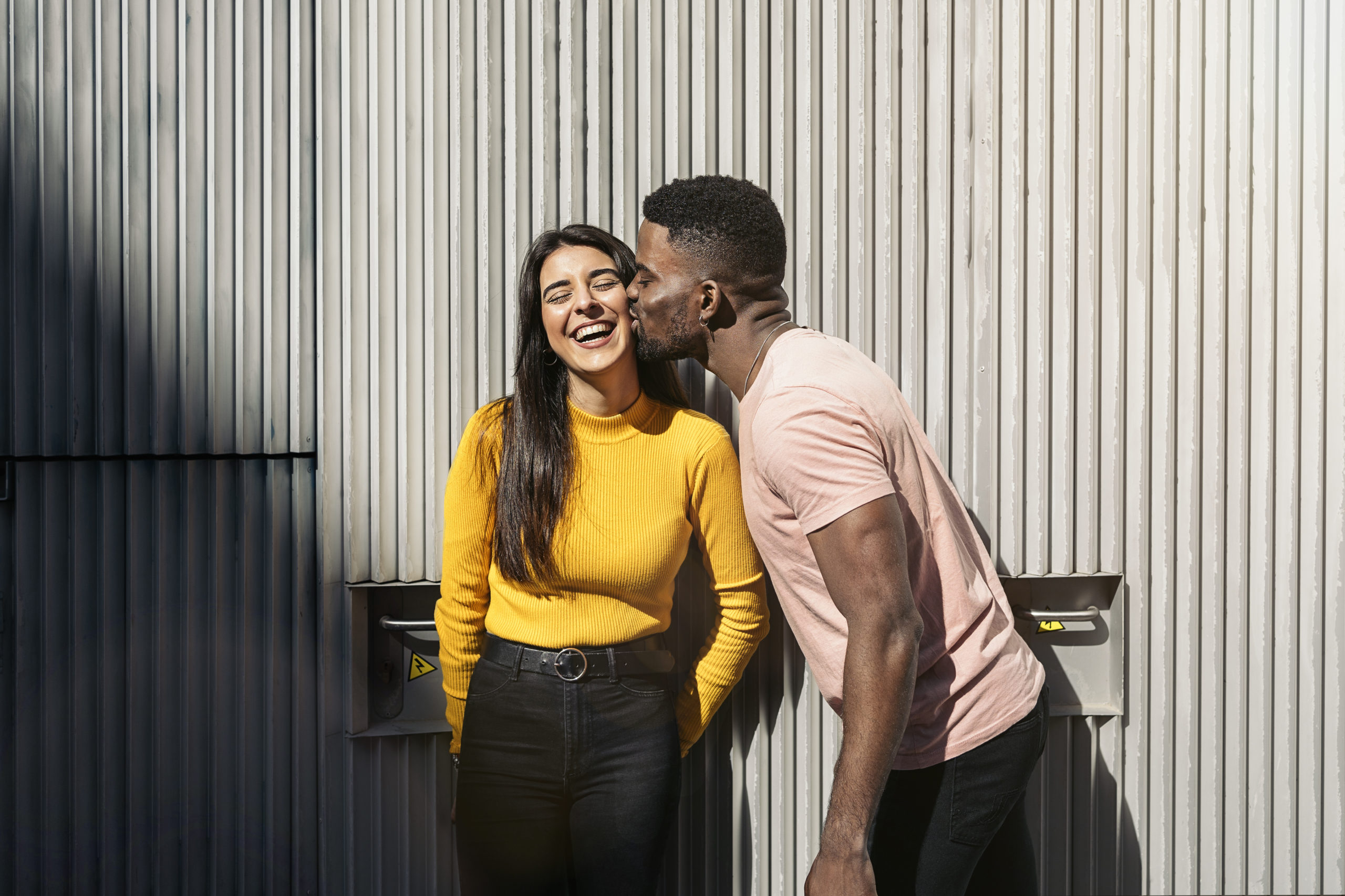 Happy couple standing outdoors with man kissing woman on the cheek as she smiles and asks herself the question, "is he serious about me"?