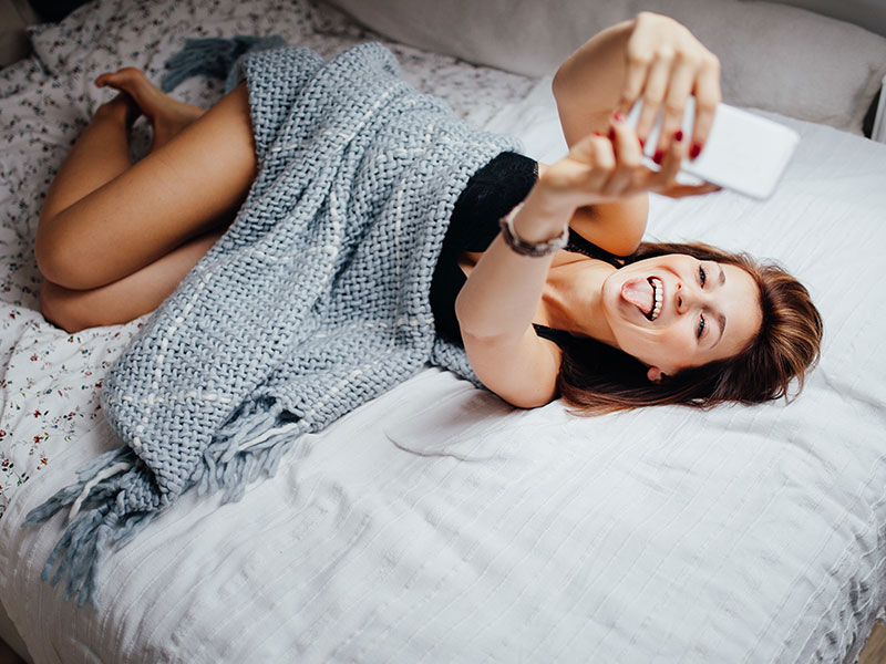 A girl text flirting laying on her bed and sticking her tongue out at her phone to send a flirty text.