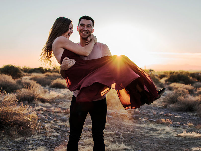 A man who was wondering does she love me, holding is girlfriend in his arms on the beach while she laughs.