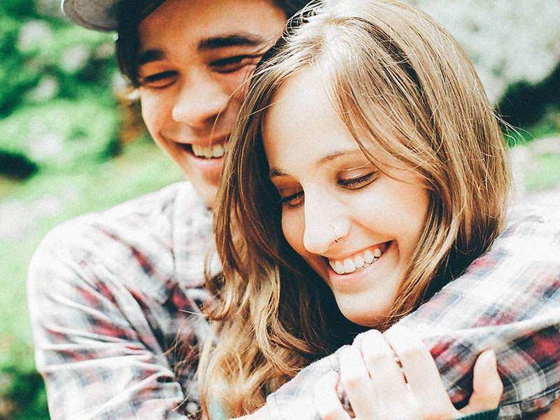 A clingy girlfriend hanging onto her boyfriend while they kiss and laugh outside.