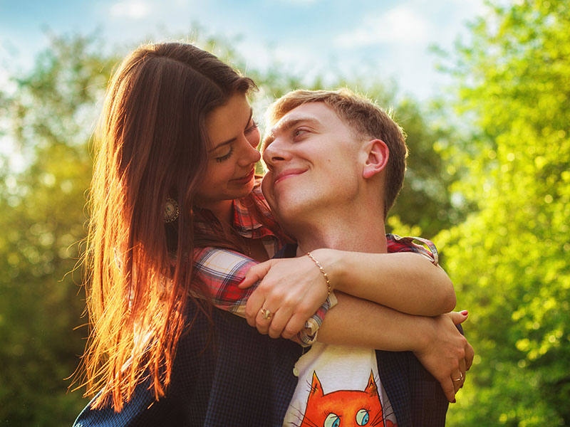 A woman who learned how to make the first move on a guy, hugging a guy from behind and flirting while he smiles.