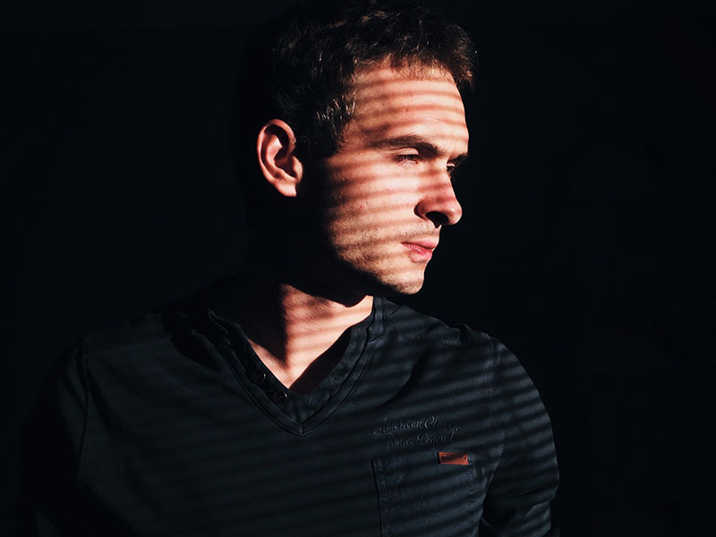 A man who’s dating a narcissist looking out the window with the blinds casting a shadow on him as he thinks about his relationship.