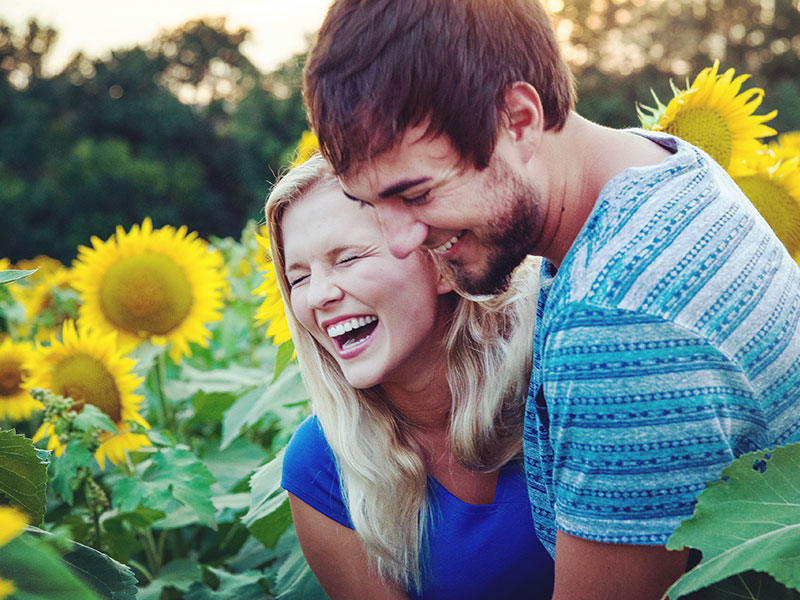 A couple in a honeymoon period laughing in a field of sunflowers together.