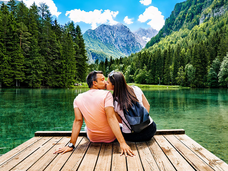 A woman dating an older man, kissing him on a dock in front of a beautiful mountain.
