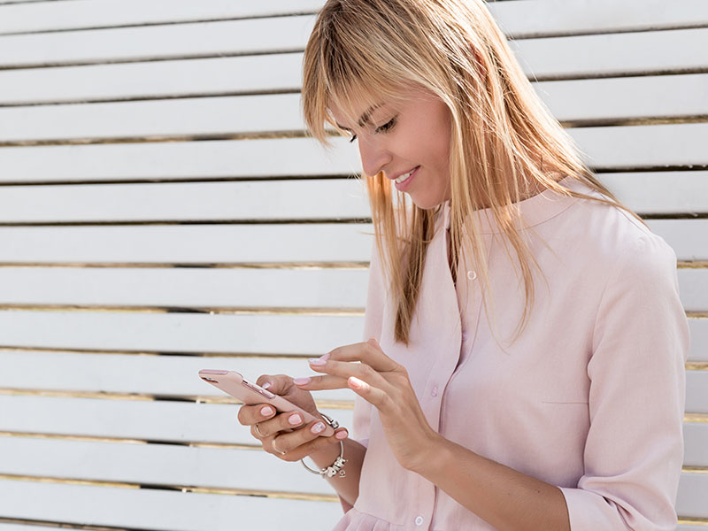 A woman texting a guy who knows how to text a girl back, smiling and looking happy in the sun.