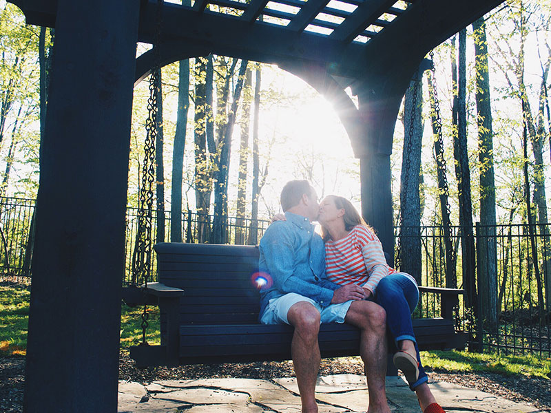 A couple who met on over 50 dating sites, kissing on a park bench as the sun shines through the trees.