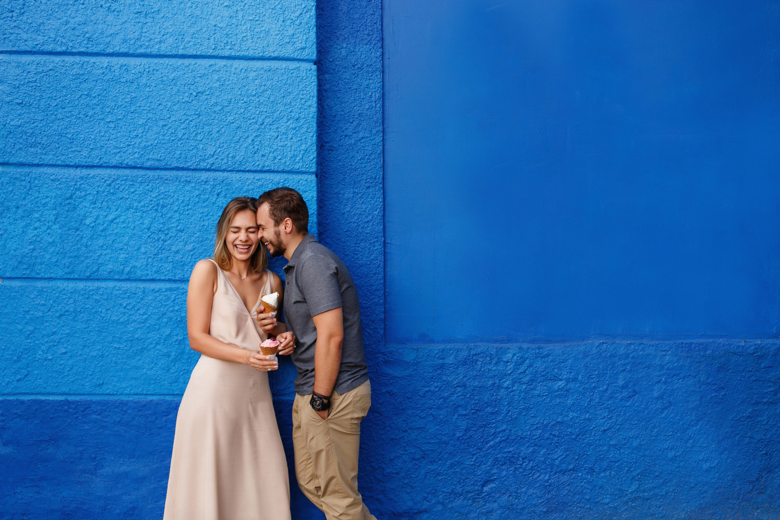 Happy, laughing man and woman standing together in front of a blue-painted wall while the woman tries out some flirty questions to ask a guy.