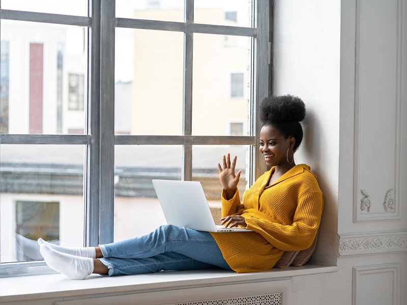 woman sitting on window sill with laptop waving at long distance partner on Zoom call