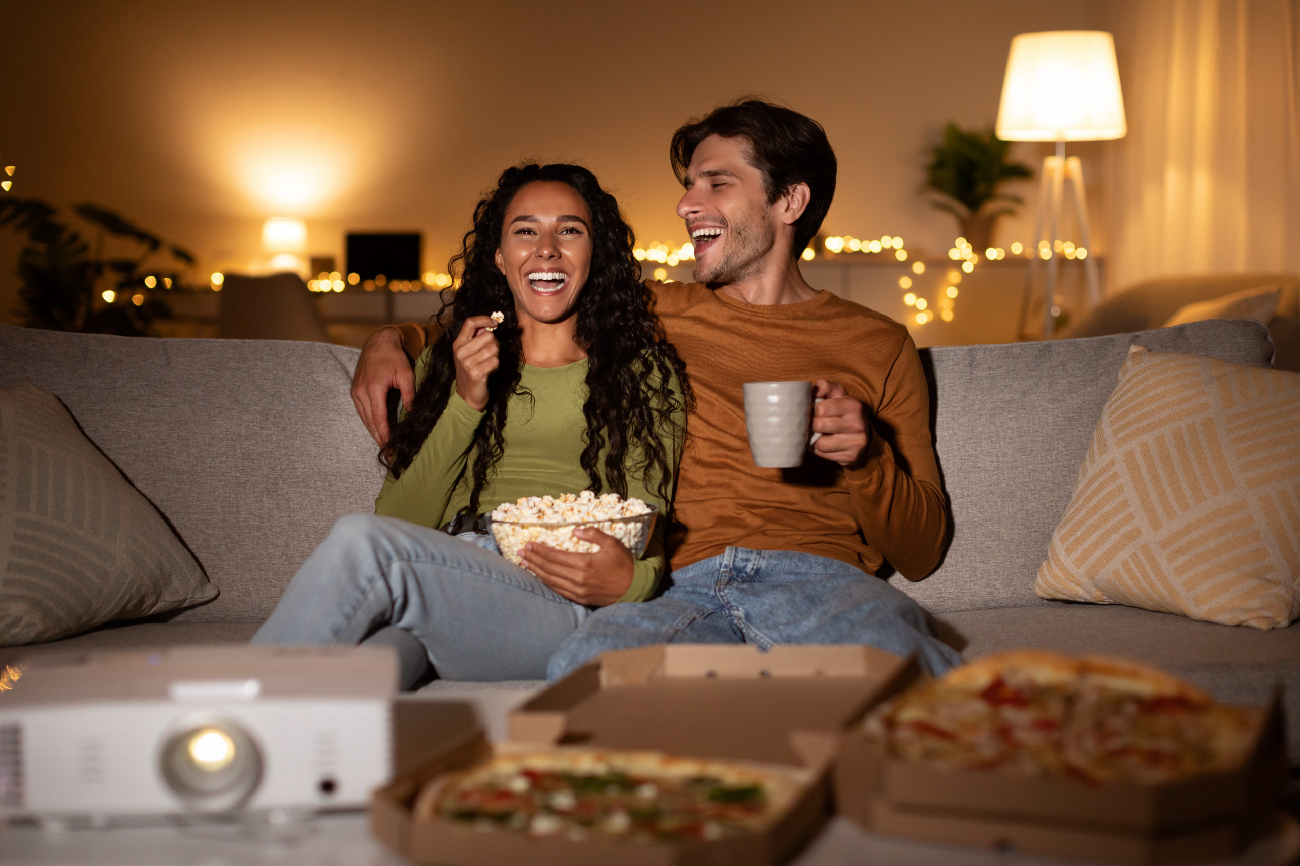 Happy couple sitting on sofa together and laughing while trying out some romantic things to do at home.