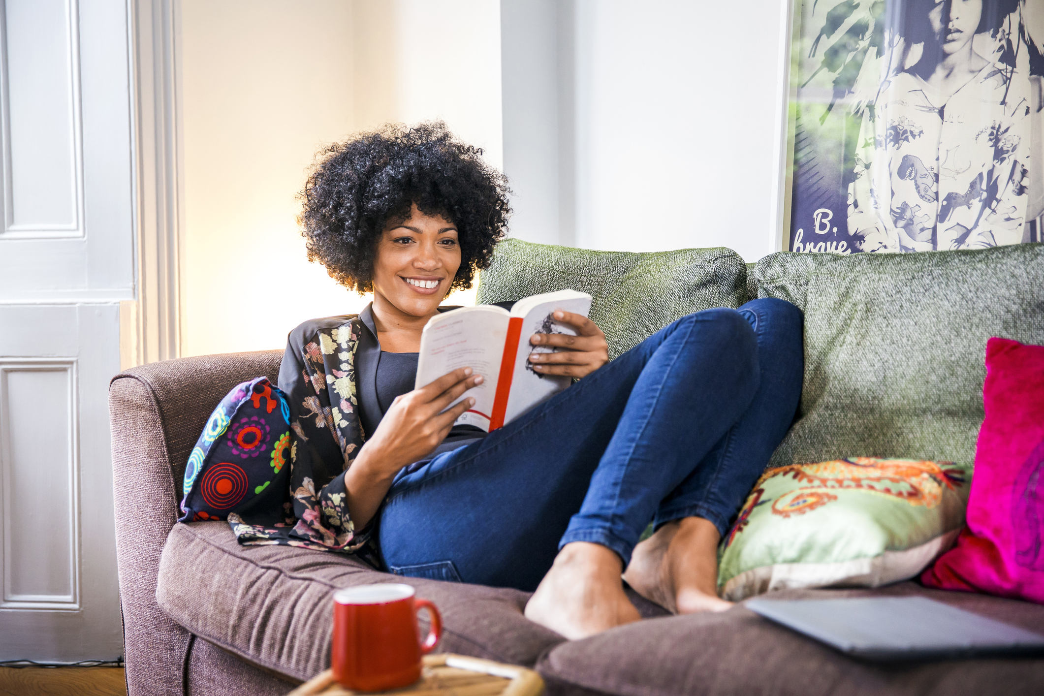 Happy, millennial woman sitting on the couch and smiling while reading one of the best romance novels of all time.