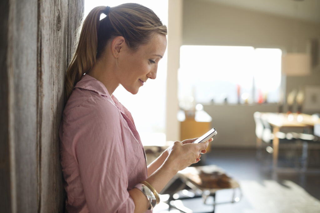Smiling woman standing in house, leaning against the wall and using mobile phone for online dating in Sacramento.