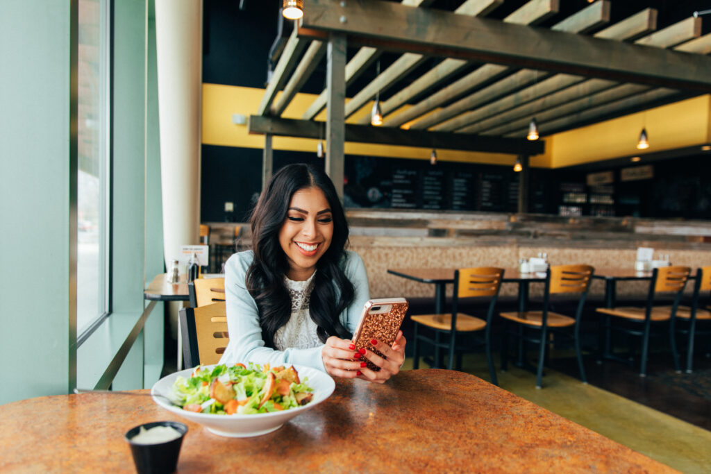Happy American-Mexican woman sitting in restaurant and smiling while using her mobile phone for Mexican dating.